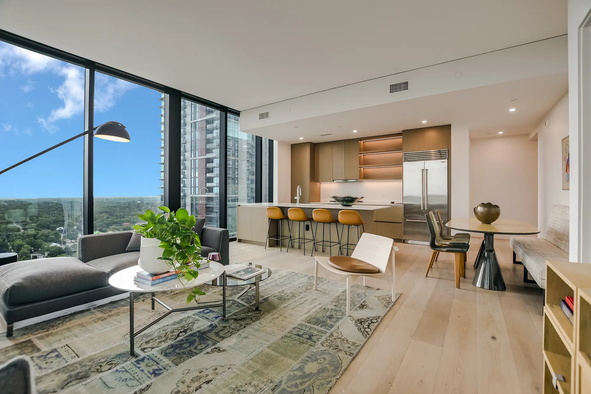 천장까지 닿는 창문으로 도시 경관이 보이는 모던한 거실. 아늑한 좌석 공간, 바 스툴이 있는 오픈형 주방, 밝은 색상의 나무 바닥이 특징입니다.