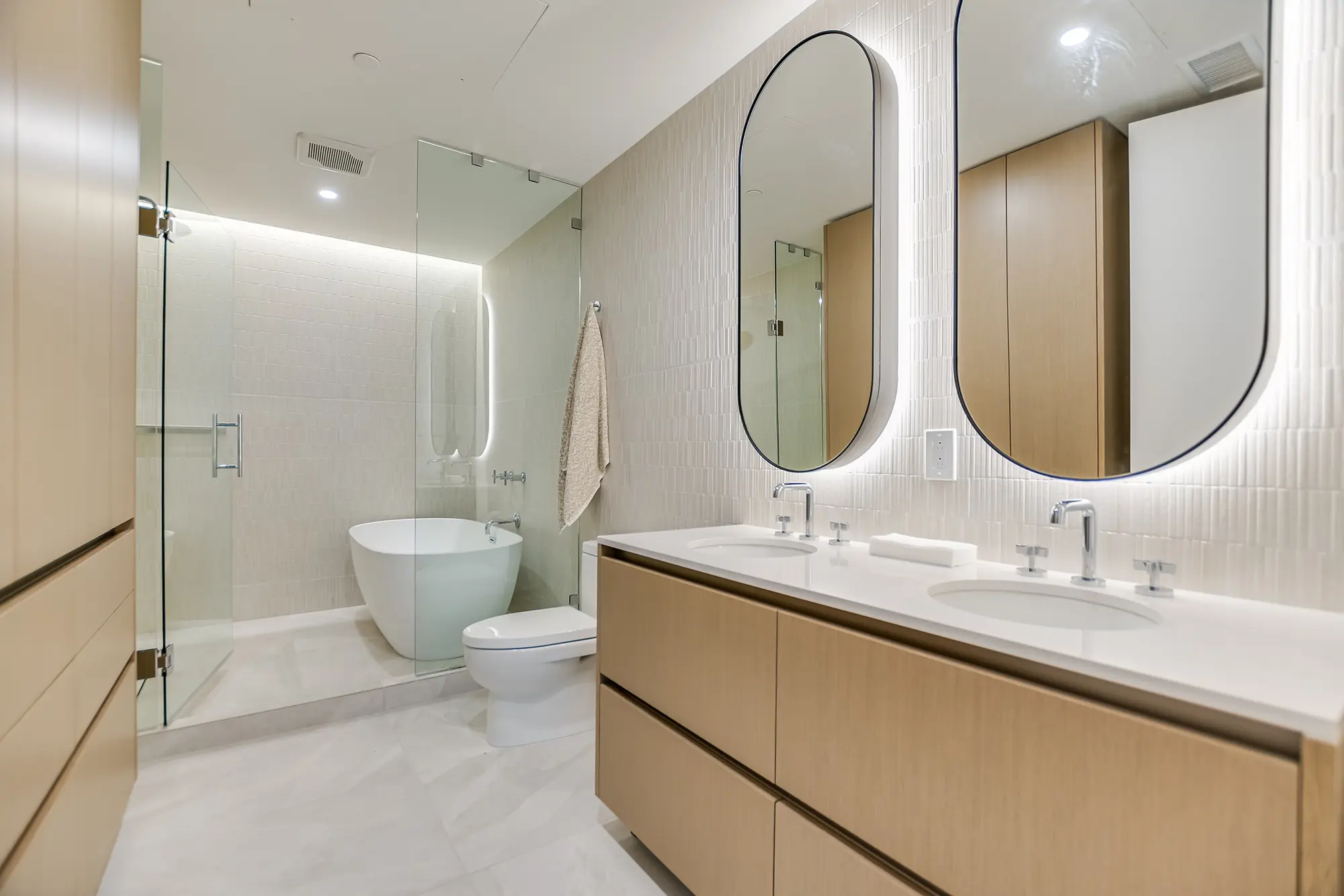 현대적인 욕실에는 목재 캐비닛, 타원형 거울이 달린 더블 세면대, 그리고 독립형 욕조가 있는 유리로 둘러싸인 샤워 공간이 있습니다. 깔끔하고 미니멀한 디자인입니다.