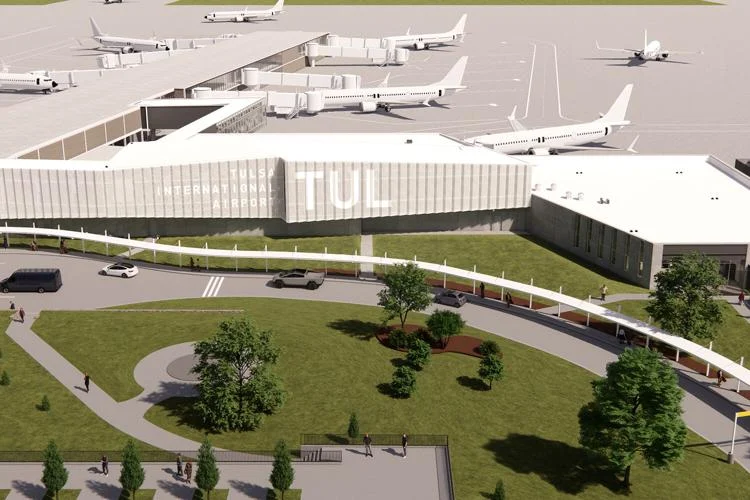 Wide view of Tulsa International Airport showing modern terminal building labeled "TUL," surrounded by parked vehicles, green trees, and several airplanes on the tarmac.