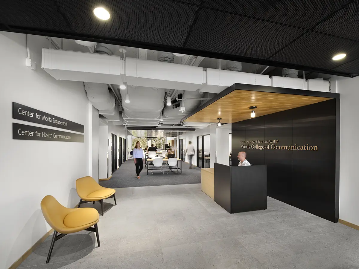 Modern office lobby with minimalist decor at Moody College of Communication. Features include yellow chairs, wall signs, and individuals engaged in conversation.