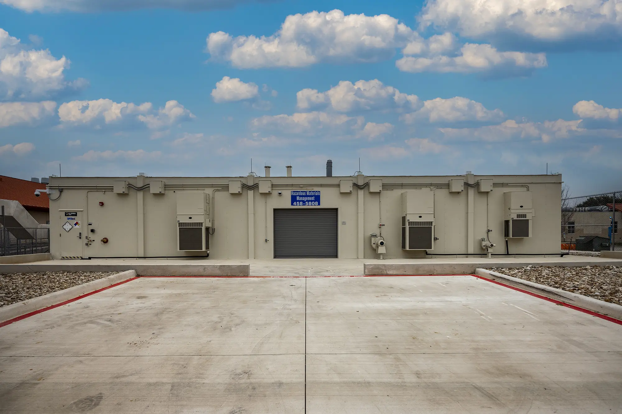 Flat-roofed beige building with utility boxes and a garage door, under a blue sky with fluffy clouds. Concrete foreground, with a calm, industrial vibe.
