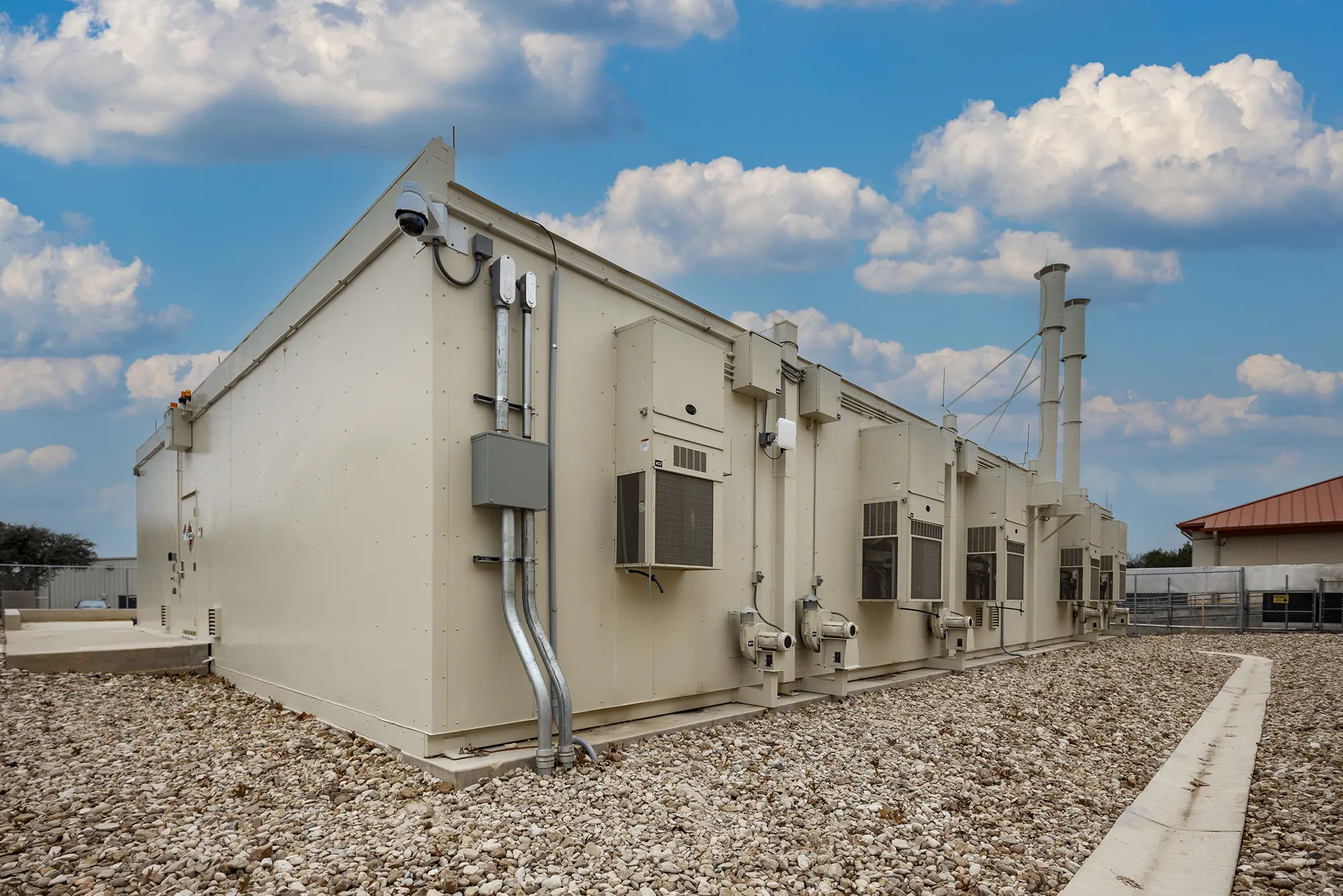 Large beige industrial building with multiple air conditioning units and vents, set on a gravel surface. The sky is blue with scattered clouds.