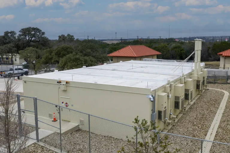 A large, beige building with a flat white roof, surrounded by a chain-link fence. Air conditioning units line the side. Trees and red-roofed structures are in the background.