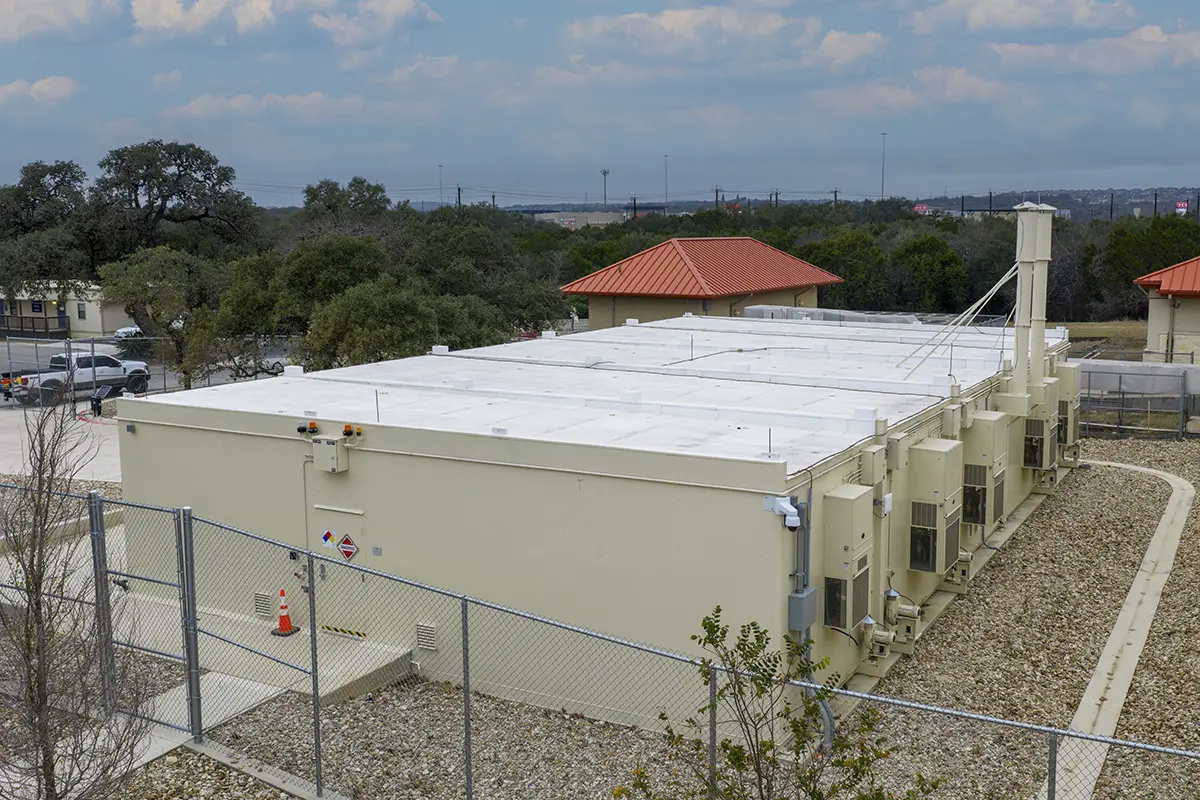 A large, beige building with a flat white roof, surrounded by a chain-link fence. Air conditioning units line the side. Trees and red-roofed structures are in the background.