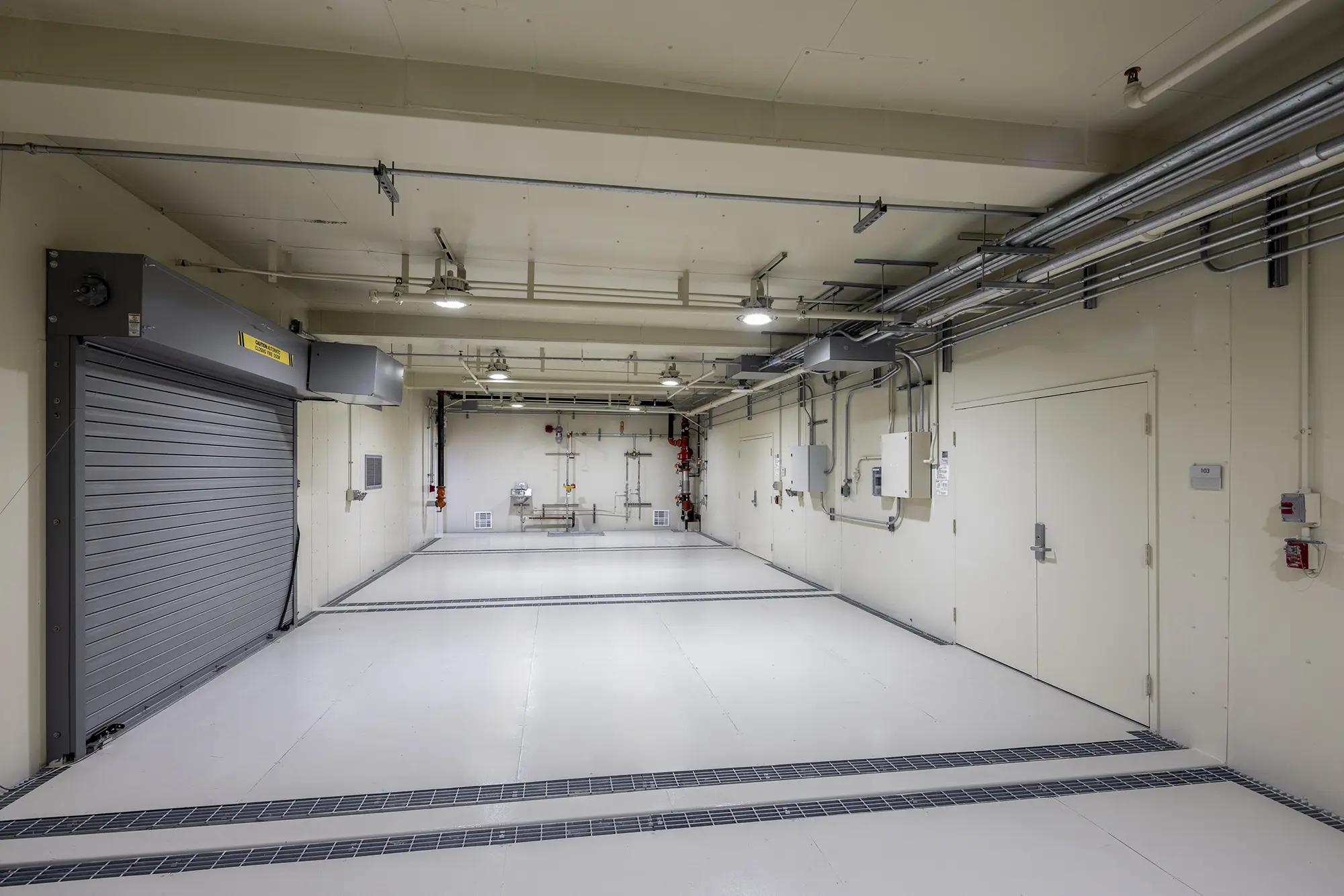 Industrial room with a clean, sterile environment. Features a large metal roller door, white walls, ceiling pipes, and evenly spaced lighting.