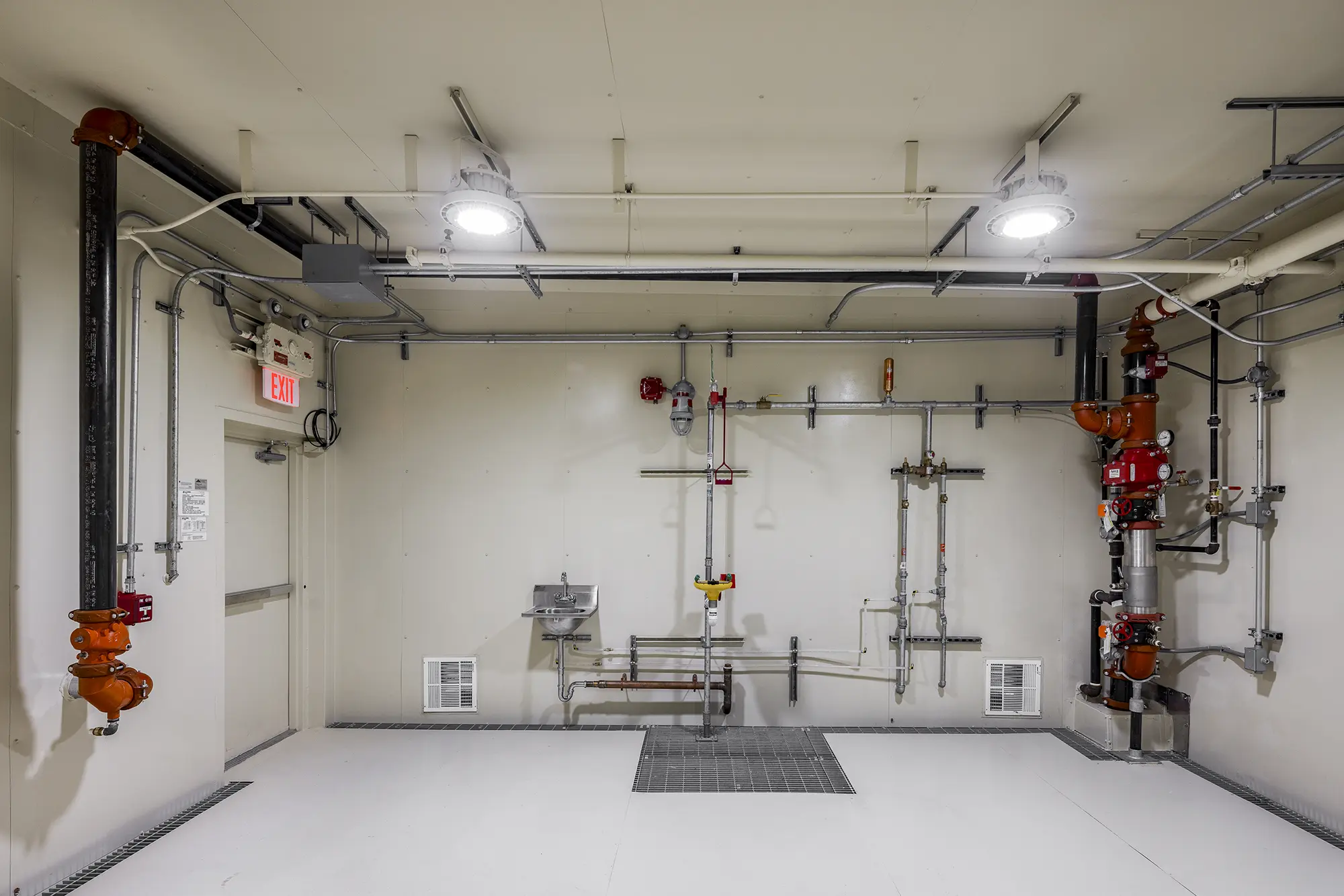 Industrial room with exposed pipes and valves against a white wall. Bright overhead lights illuminate a metal door with an exit sign and a small basin.