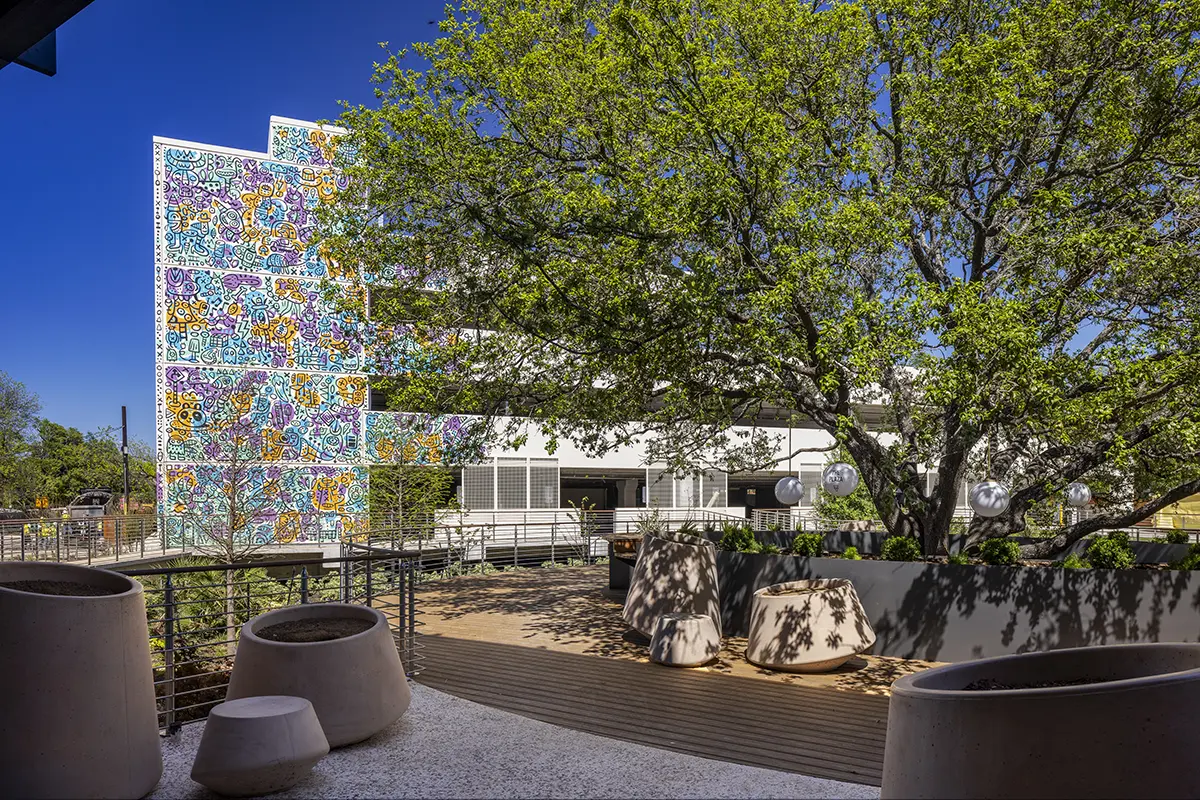 Outdoor patio with modern planters and a tree under a clear blue sky. Background features a colorful mural on a building, adding vibrancy.