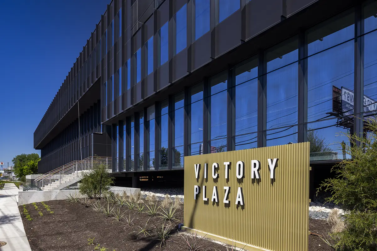 Modern building with reflective blue windows at Victory Plaza. The beige sign contrasts the sleek black facade. Sunny day, minimal landscaping.