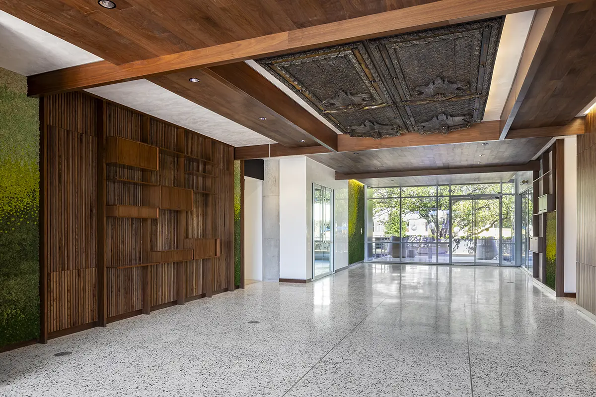 Spacious modern room with terrazzo floor, wooden ceiling accents, and green wall elements. Large glass windows offer natural light and outdoor view.