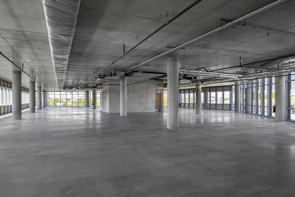 Spacious empty warehouse with exposed concrete floors, large windows, and industrial ceiling ducts. Natural light creates a bright, open atmosphere.