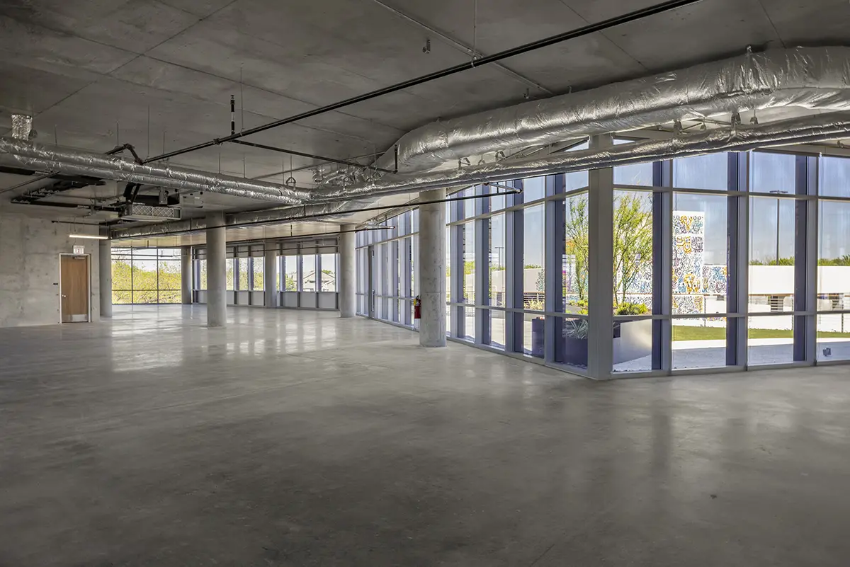 Spacious empty office with concrete floors, exposed ductwork, and large floor-to-ceiling windows offering a view of trees and buildings outside. Modern and industrial.