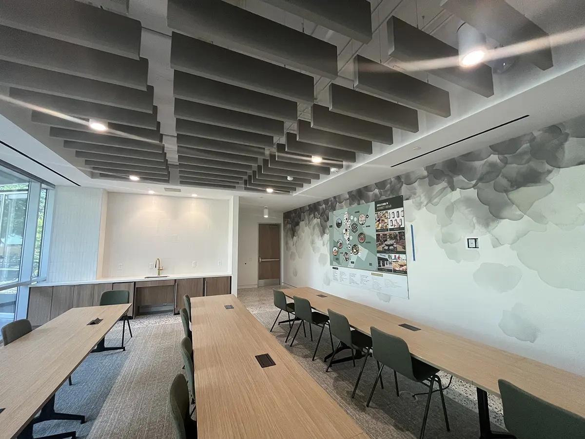 Modern meeting room with light wooden tables and green chairs. Acoustic panels hang from the ceiling. A mural covers one wall, adding a creative touch. Soft, neutral colors create a calm atmosphere.