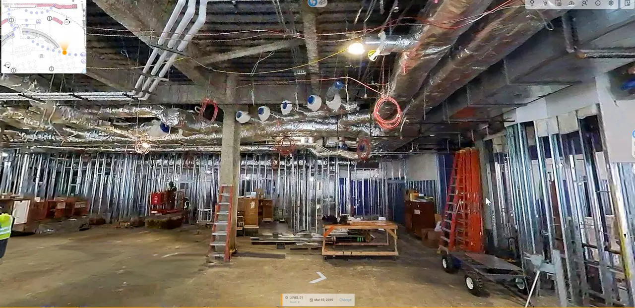 Interior construction site with exposed ductwork, metal framing, ladders, and scattered tools. A worker in a safety vest stands on the left. Industrial tone.
