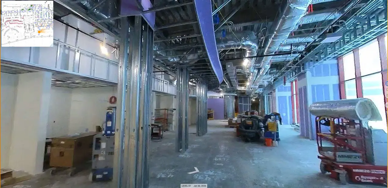 Construction site interior with exposed metal beams, ducts, and scattered equipment. Large windows on the right let in daylight. Active work area.
