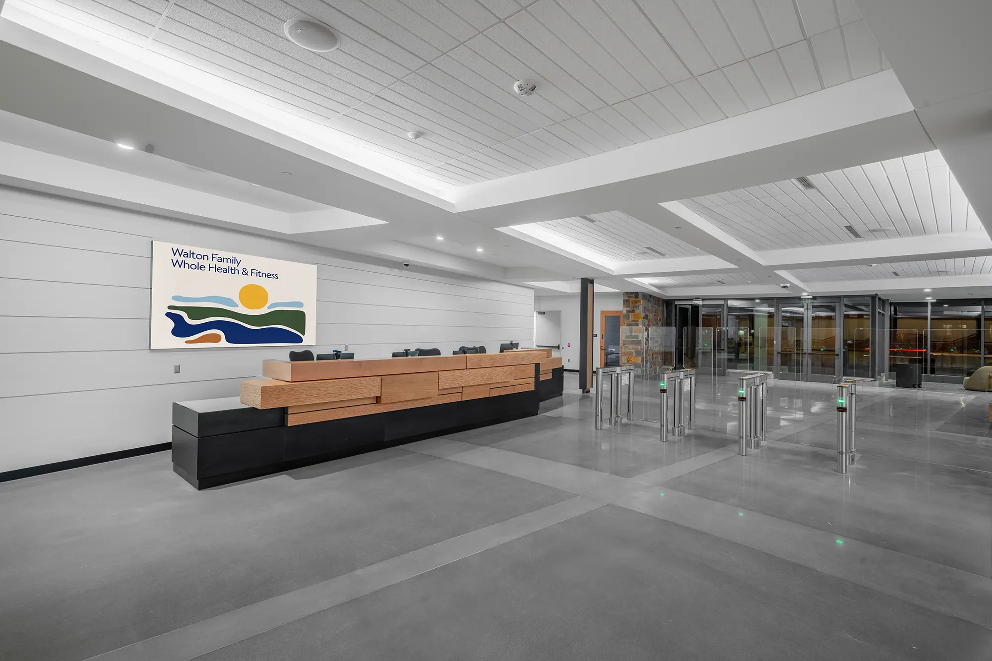 Spacious, modern reception area of a fitness center with a sleek wooden desk, labeled sign on the wall, and metallic turnstiles for entry.