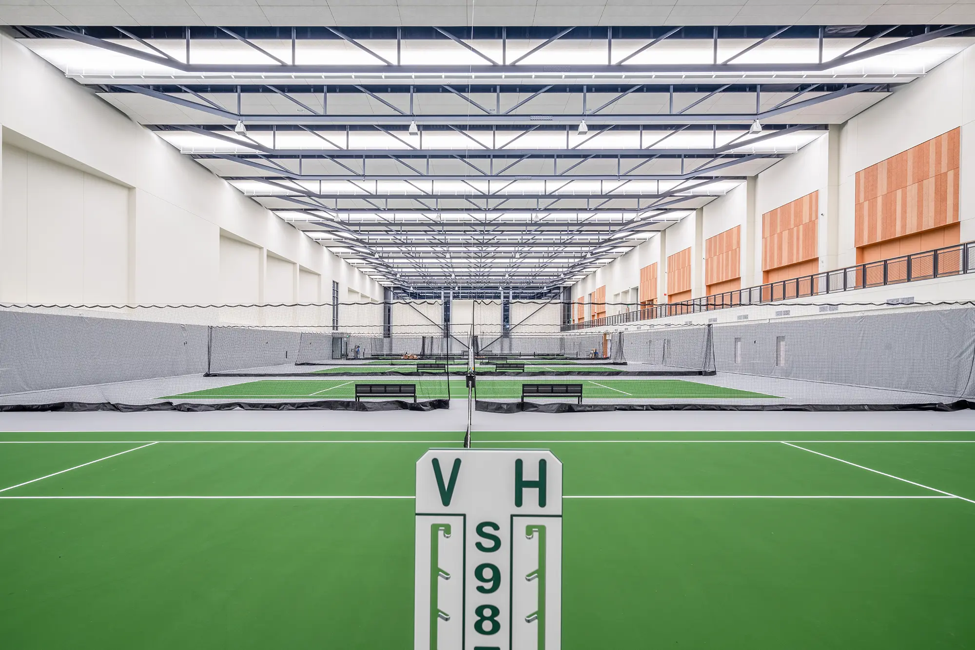 Indoor tennis court with vibrant green flooring, surrounded by high walls and an airy ceiling. A scorecard stands in the foreground, enhancing the competitive feel.