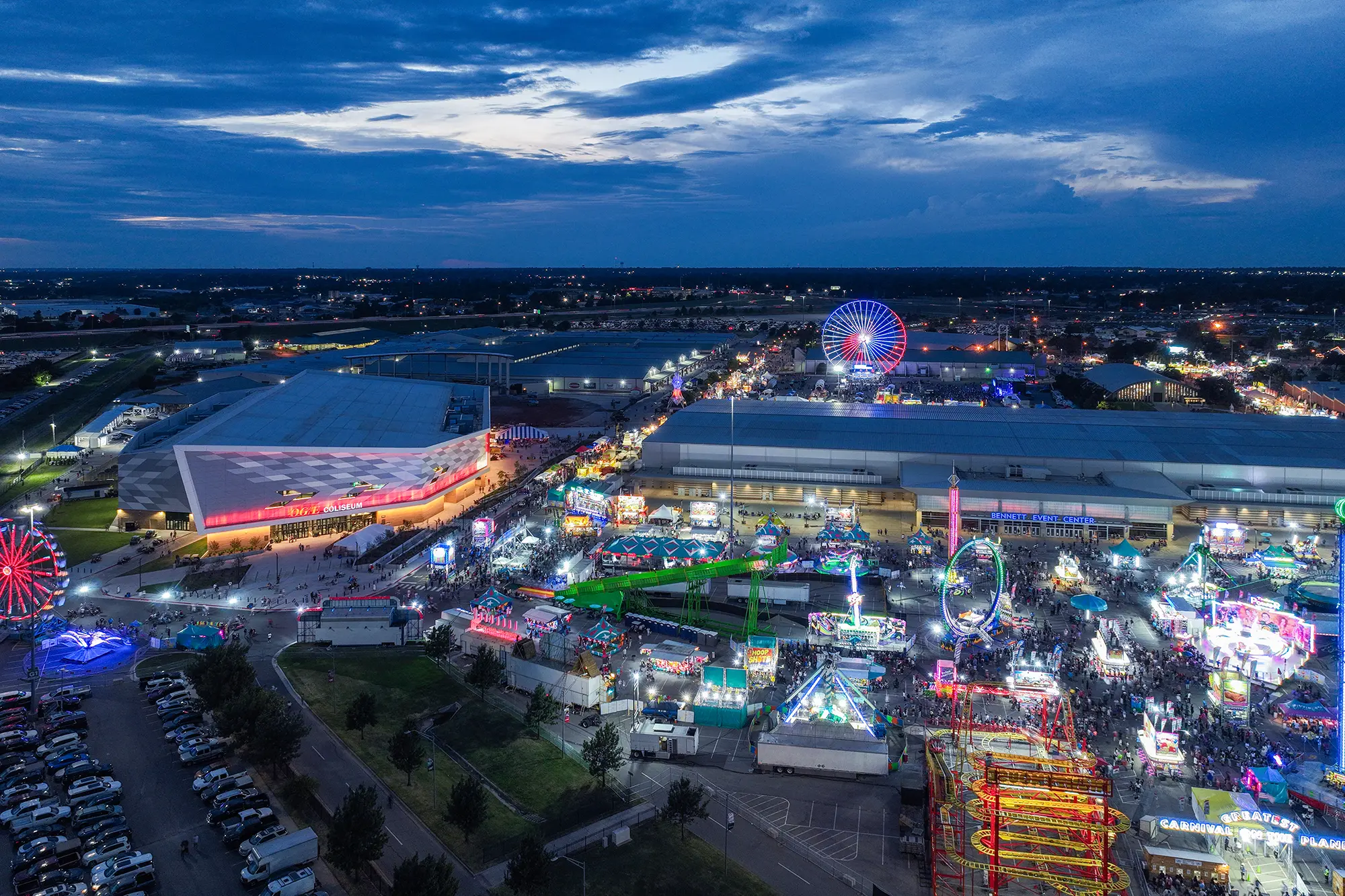 A vibrant aerial view of a lively fairground at dusk with colorful rides and glowing lights. People crowd the area, creating a festive atmosphere under a dramatic, partly cloudy sky.
