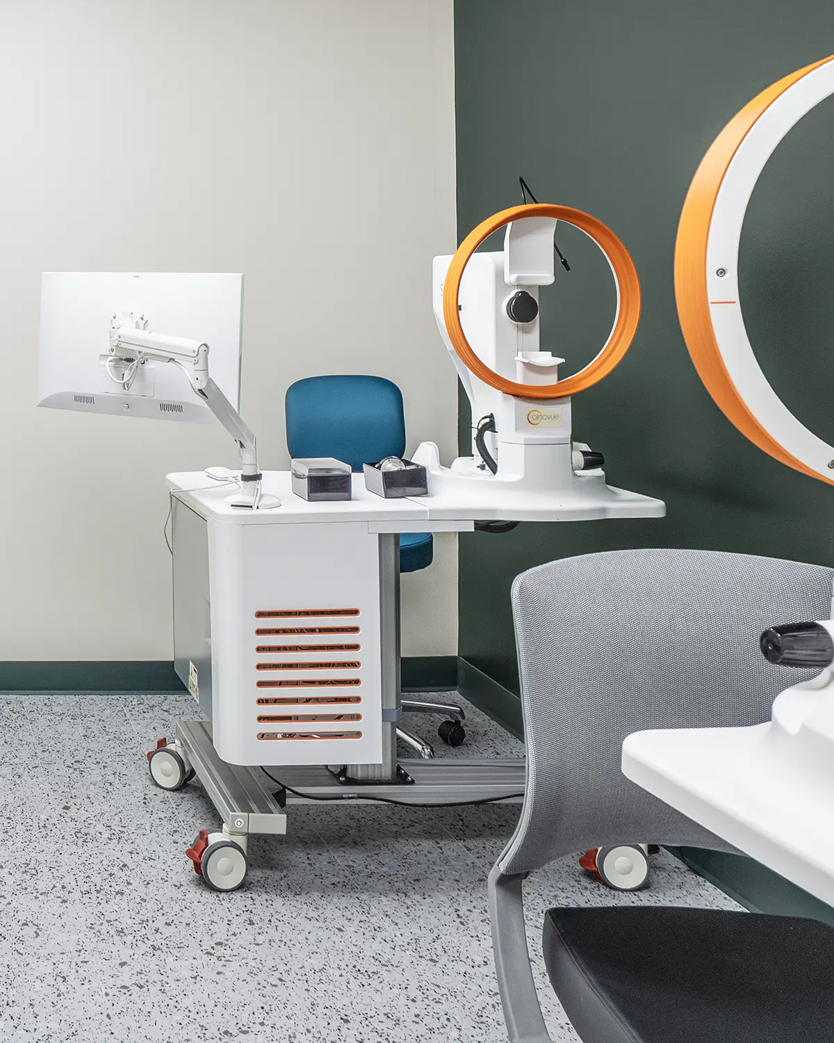 Modern medical examination room featuring advanced ophthalmology equipment with orange accents, a blue chair, and clean, minimalist design.