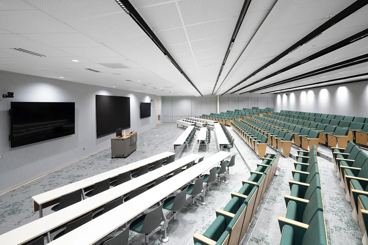 A modern lecture hall with tiered seating in green, sleek white tables, and multiple large screens on gray walls. Bright lighting and a clean, orderly atmosphere.