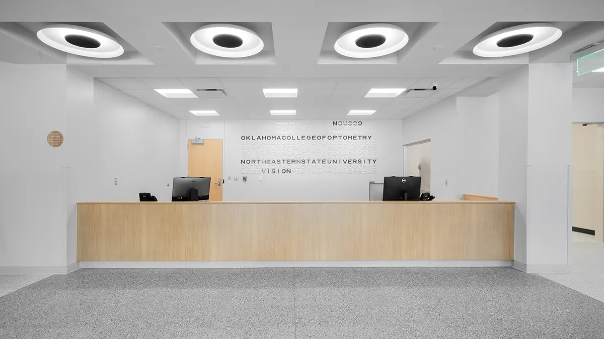 Modern reception area with a light wood desk, two computers, and minimalistic decor. Circular ceiling lights cast soft, diffused light. Walls are white.