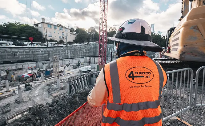 A construction worker in an orange safety vest and helmet overlooks a large construction site with heavy machinery and building materials, conveying a sense of industry and focus.