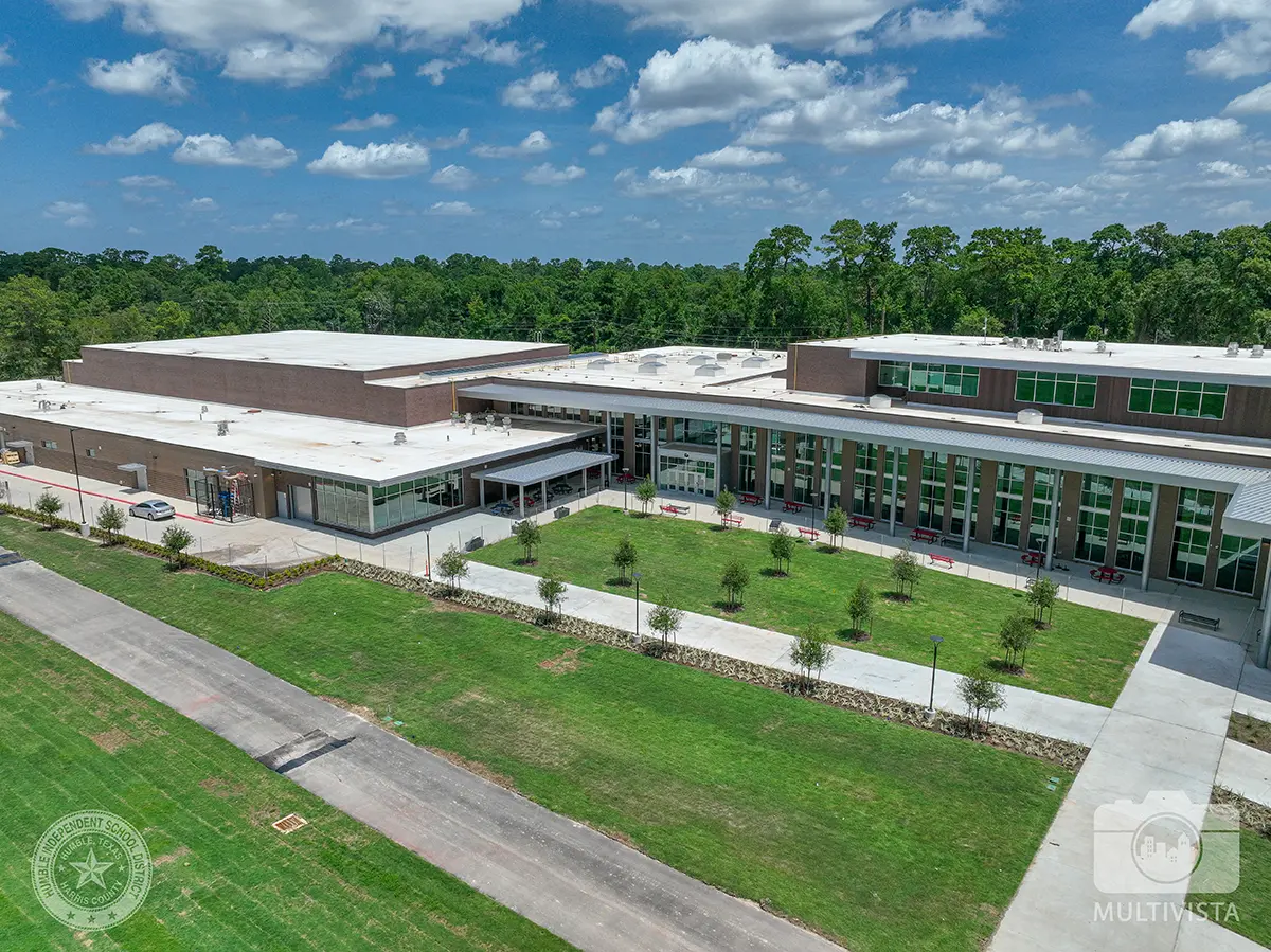 Aerial view of a modern, two-story brick school building surrounded by lush green lawns and trees under a partly cloudy sky, creating a serene atmosphere.