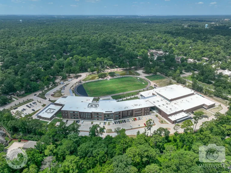 Aerial view of a school campus surrounded by dense forest. The main building has a white roof, with a sports field and track behind it. The setting is peaceful and expansive.